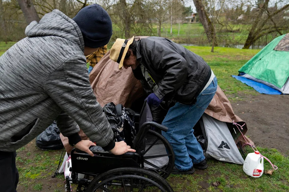 A person wearing a dark sweatshirt and hat holds onto a wheelchair as a man crouches in front of a tent next to the wheelchair.