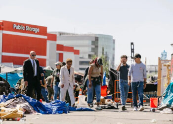 A woman wearing a white pantsuit walks alongside a few people in a street that is littered in front of a few large buildings.