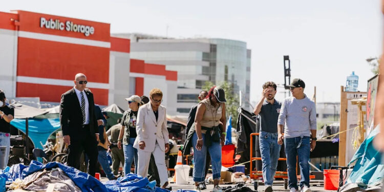 A woman wearing a white pantsuit walks alongside a few people in a street that is littered in front of a few large buildings.