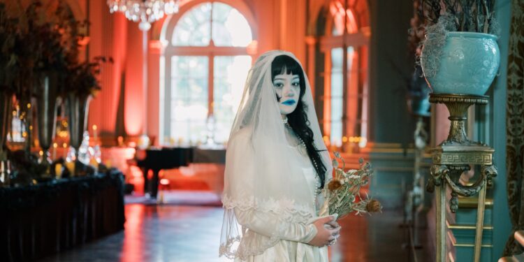 A woman wearing a white dress, veil and ghostly make-up stands in a grand hall, lit in red. She is holding dried flowers.