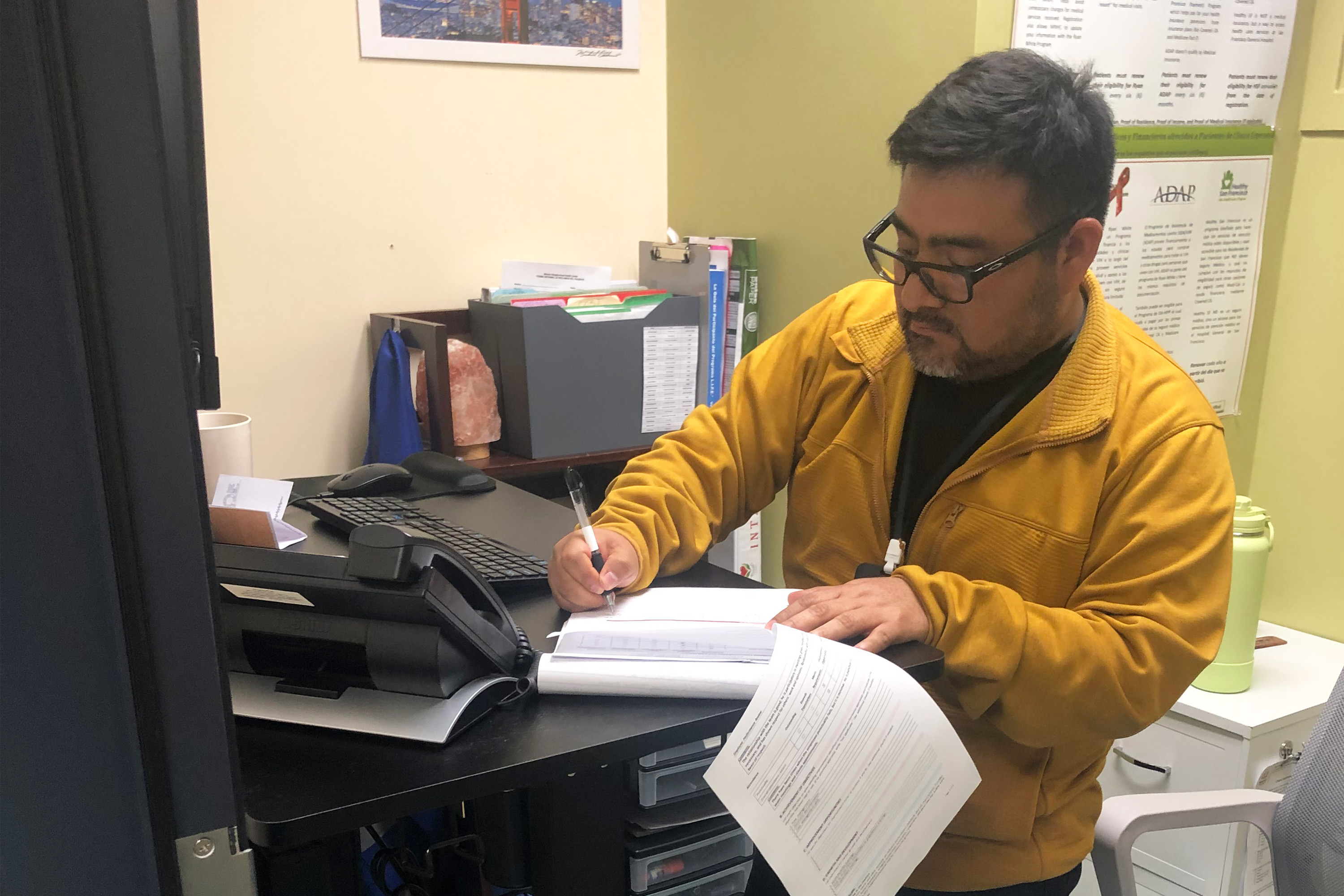 A photo of a man standing by a desk, holding papers.