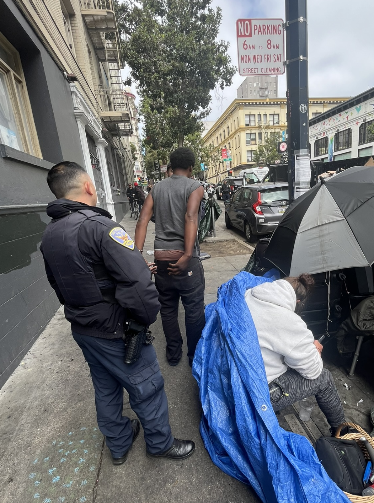 San Francisco police officers detain a man for 'illegal lodging' under a tarp and umbrella in the Tenderloin.